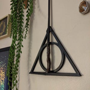 May include: A black metal wall mount in the shape of the Deathly Hallows symbol from the Harry Potter series. A black wand is hanging from the mount.