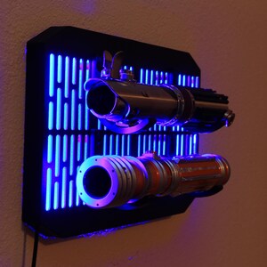 May include: A black wall mount with a blue light display. Two lightsabers are mounted on the wall mount. The lightsabers are silver and orange. The wall mount is shaped like a grid with vertical lines.