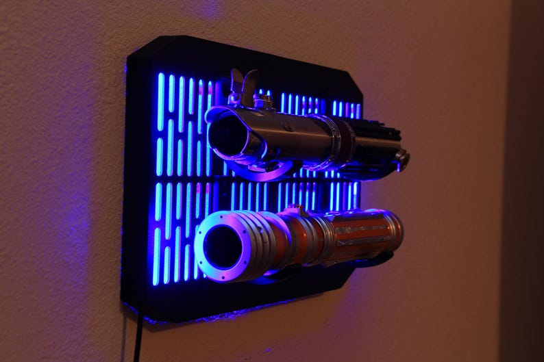May include: A black wall mount with a blue light display. Two lightsabers are mounted on the wall mount. The lightsabers are silver and orange. The wall mount is shaped like a grid with vertical lines.