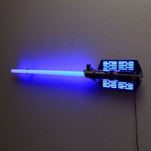 Puede incluir: Decoraci&oacute;n de pared de sable de luz azul con un soporte de montaje negro. La hoja del sable de luz brilla con una luz azul brillante. El soporte tiene una serie de luces azules. Un cable de alimentaci&oacute;n negro se extiende desde el soporte.