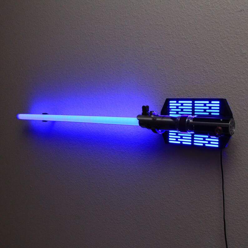 Puede incluir: Decoraci&oacute;n de pared de sable de luz azul con un soporte de montaje negro. La hoja del sable de luz brilla con una luz azul brillante. El soporte tiene una serie de luces azules. Un cable de alimentaci&oacute;n negro se extiende desde el soporte.