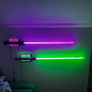 Puede incluir: Dos luces montadas en la pared con forma de sables de luz. La superior brilla en p&uacute;rpura, la inferior en verde. Los mangos son negros y grises. Las luces est&aacute;n montadas en una pared gris.