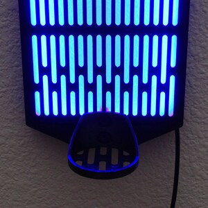 May include: A black wall mount with a blue light grid pattern and a black plastic holder underneath. The holder has a grid pattern and is designed to hold a phone or other device.