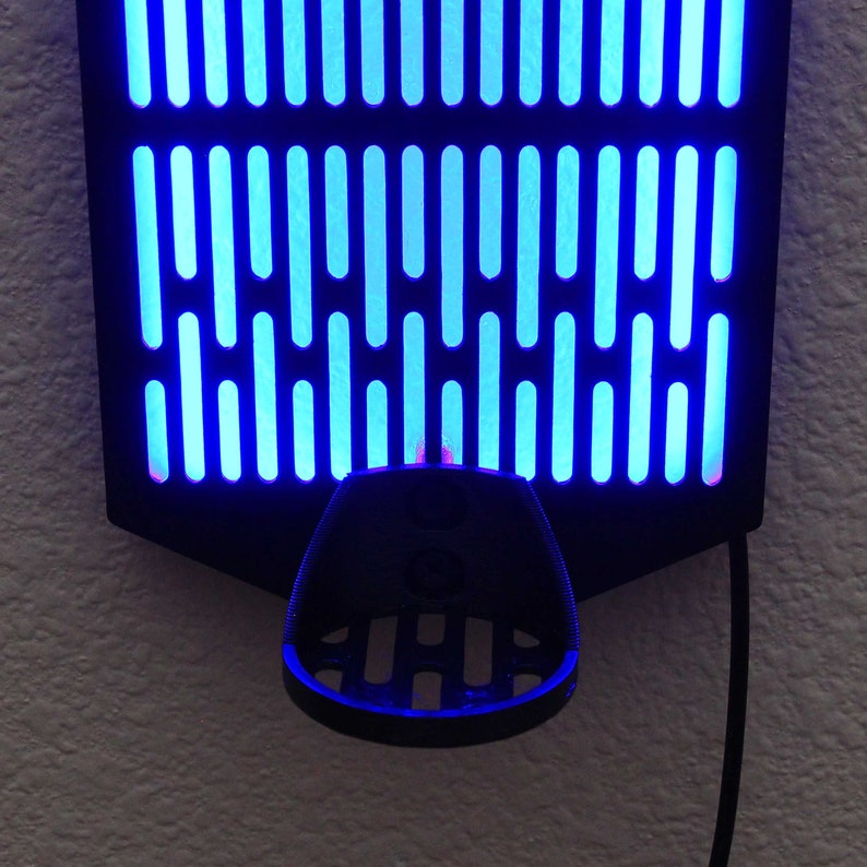 May include: A black wall mount with a blue light grid pattern and a black plastic holder underneath. The holder has a grid pattern and is designed to hold a phone or other device.