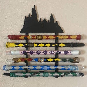 Può includere: Un supporto da parete in metallo nero a forma di castello di Hogwarts con sei bacchette magiche esposte. Le bacchette sono dipinte in vari colori e motivi, tra cui giallo, rosso, blu, verde e rosa. Alcune delle bacchette hanno cristalli all'estremità.