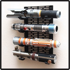 Puede incluir: Cuatro accesorios de sable de luz montados en un soporte de pared negro. Los sables de luz son plateados, azules, naranjas y negros con detalles marrones.