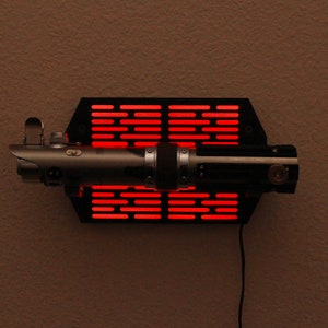Puede incluir: R&eacute;plica de sable de luz plateada y negra montada sobre una base hexagonal negra con iluminaci&oacute;n LED roja. El sable de luz tiene una empu&ntilde;adura detallada y se exhibe horizontalmente contra una pared de color neutro. Un cable de alimentaci&oacute;n sale de la base.