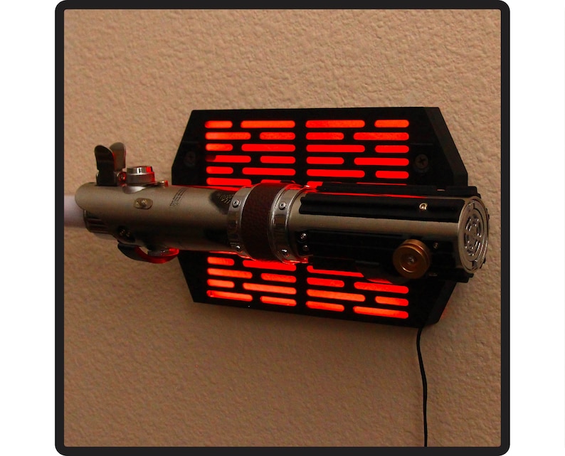 Puede incluir: Un sable de luz plateado y negro montado en una pantalla de pared negra con detalles rojos brillantes. El sable de luz est&aacute; detallado con varios componentes met&aacute;licos y marrones, y est&aacute; conectado a una fuente de alimentaci&oacute;n.