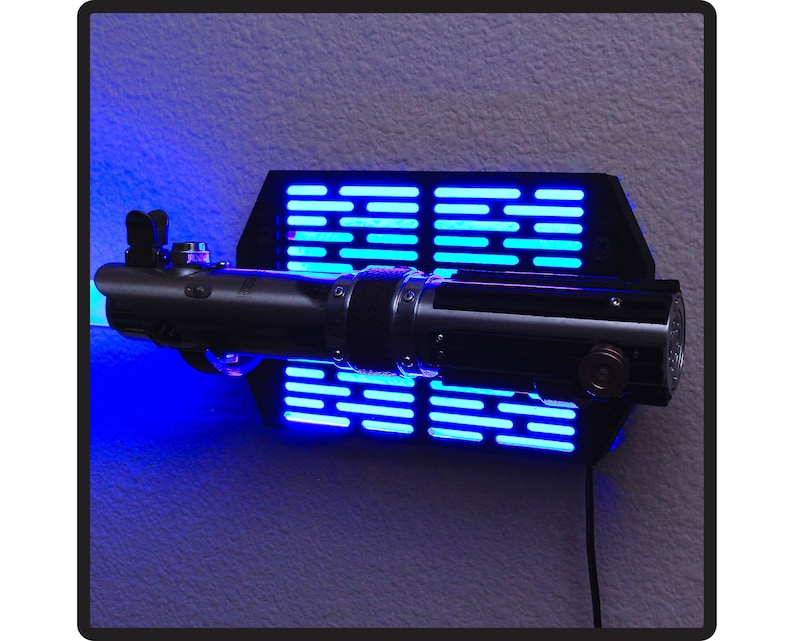 Puede incluir: Un sable de luz negro y plateado montado en una pantalla de pared negra con luces LED azules. El sable de luz est&aacute; detallado con detalles met&aacute;licos y un dise&ntilde;o cil&iacute;ndrico. La pantalla tiene forma rectangular con una serie de luces azules horizontales.