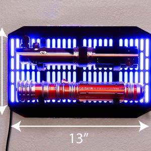 May include: A black lightsaber wall mount with a blue LED light strip. The mount has a grid pattern and is designed to hold two lightsabers. The mount is 13 inches wide and 8 inches tall. The mount is holding two lightsabers, one red and one black.
