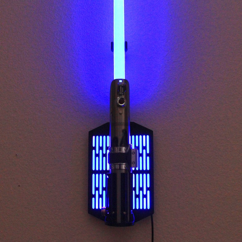 May include: A blue lightsaber with a black wall mount featuring a grid pattern. The lightsaber is lit up and the blue light is illuminating the wall behind it.
