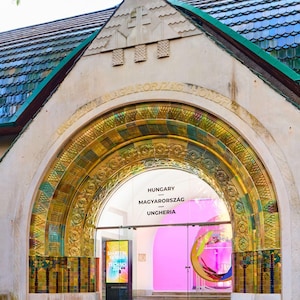 May include: A white building with a large arched entrance decorated with gold and green tiles. The archway is inscribed with the words "Hungary - Magyarorszag - Ungheria".