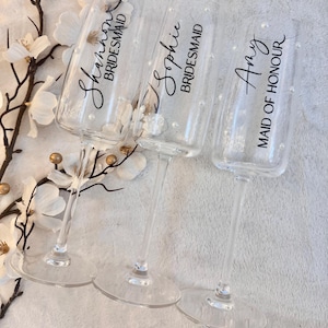 May include: Three clear champagne flutes, each with a name and title printed on them. The first reads "Shannon BRIDESMAID", the second "Sophie BRIDESMAID", and the third "Amy MAID OF HONOUR". Each glass is adorned with small, white pearl-like decorations.
