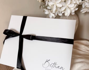 May include: A white gift box tied with a black satin ribbon. The box has the name "Bethan" and the word "Bridesmaid" printed on it in a delicate script font. A bouquet of white flowers is in the background.