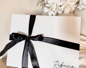 May include: A white gift box tied with a black satin ribbon and bow. The name "Rebecca" is written in cursive on the box. A vase of white flowers is in the background.