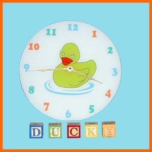 May include: A white clock with a green rubber ducky in a blue bathtub on the face. The clock has black hands and colorful numbers. The numbers are 1, 2, 3, 4, 5, 6, 7, 8, 9, 10, 11, and 12. Below the clock are five colorful blocks that spell out the word "DUCK".