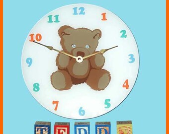 May include: A white clock with a brown teddy bear on the face. The clock has colorful numbers and hands. The numbers are 12, 1, 2, 3, 4, 5, 6, 7, 8, 9, 10, and 11. The hands are black and gold. Below the clock are five wooden blocks that spell out the word "TEDD".