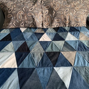 May include: A patchwork quilt made from denim fabric in shades of blue, gray, and white. The quilt is made up of triangular pieces sewn together to create a geometric pattern. The quilt is topped with two pillows with a paisley pattern in shades of brown and gray.