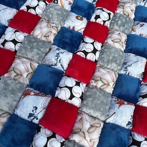May include: A handmade baseball-themed quilt featuring a patchwork design. The quilt incorporates squares of red, blue, and gray plush fabric, along with baseball-patterned fabric. Some squares feature baseball player illustrations. The quilt is designed for comfort and visual appeal.