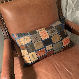May include: Rectangular denim pillow with a patchwork design of various brand labels, including "Lee," "Wrangler," and "IZOD." The pillow is placed on a brown leather armchair with a studded trim.