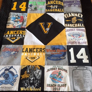 May include: A quilt made from a variety of t-shirts, each with different designs and text. The quilt is made up of 9 squares, with a large black square in the centre. The black square has a white V in the centre. The quilt is made in a variety of colours, including black, white, yellow, blue, red, and green. The text on the t-shirts includes the words "Lancers", "Baseball", "Vianney", "Jersey Shore", "Beach Blast", "North Wall", "Wave Rider", "Tournament", "12th Annual", "High School", "Best Show", "Summer Events", "Monroe Twp", "All Stars", "14", and "2012".