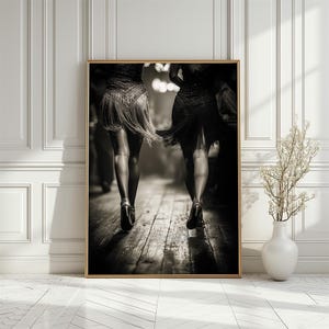 May include: Black and white framed print of two dancers in fringed dresses and high heels, viewed from the back. The dancers are on a wooden dance floor, with a blurred background. The print is in a gold frame.