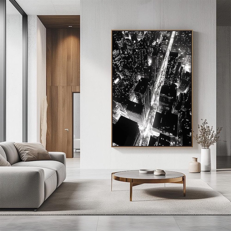 May include: Black and white aerial photograph of a city at night. The photo shows a grid of streets with buildings and lights. The photo is framed and hanging on a wall in a modern living room.
