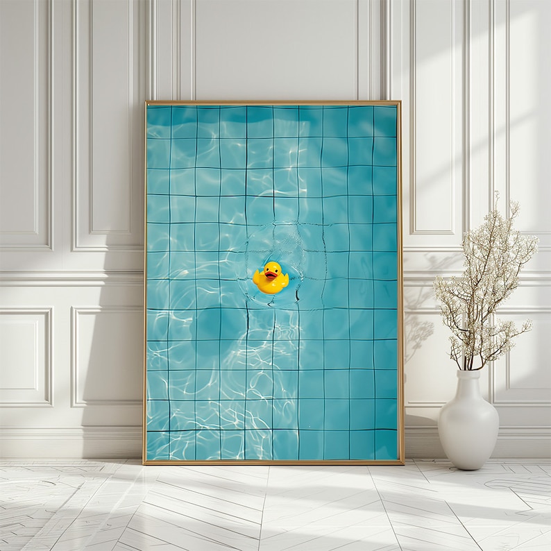 May include: A framed art print featuring a yellow rubber duck floating in a turquoise blue pool. The pool has a grid pattern, and the water reflects light. The frame is gold, and the background is a white wall.