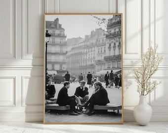 Può includere: Fotografia incorniciata in bianco e nero di una scena urbana europea. Diverse persone sono sedute su una panchina, in conversazione. Lo sfondo presenta edifici, pedoni e un'auto d'epoca, creando un'estetica classica e senza tempo.