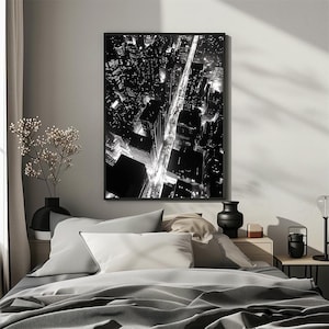 May include: Black and white photograph of a city skyline at night. The photo shows a grid of streets with buildings and lights. The photo is framed and hanging on a wall above a bed.