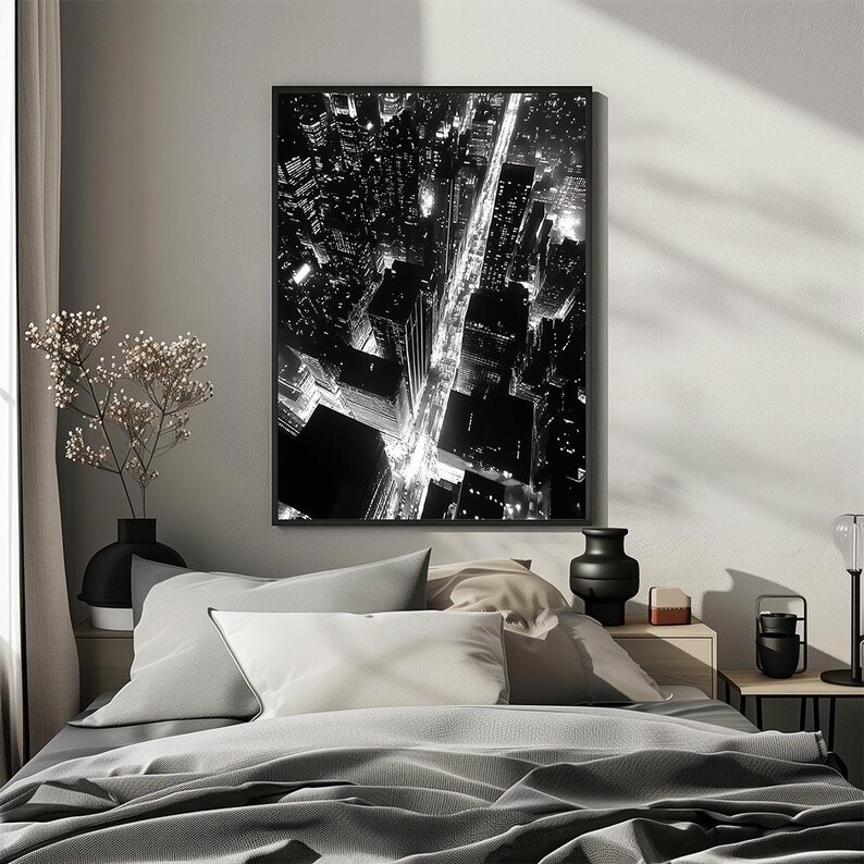 May include: Black and white photograph of a city skyline at night. The photo shows a grid of streets with buildings and lights. The photo is framed and hanging on a wall above a bed.