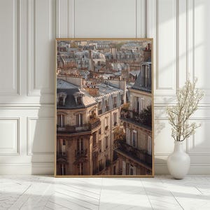 Op de afbeelding: Een ingelijste print met een stadsgezicht van Parijs, met de nadruk op de daken en architectonische details. De afbeelding toont gebouwen met lichtgekleurde gevels, dakkapellen en balkons. Het kunstwerk wordt gepresenteerd tegen een witte muur met een vaas met droogbloemen.