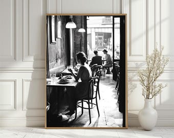 May include: Black and white photograph of a woman sitting at a table in a Parisian cafe, eating a meal. Two other people are seated at a table in the background.