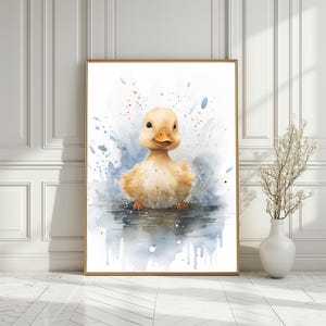 Op de afbeelding: Een aquarel schilderij van een geel eendje in een plas water. Het eendje kijkt de kijker aan met een gelukkige uitdrukking. De achtergrond is een lichtblauwe aquarel was met spatten van gouden verf.