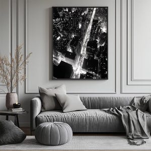 May include: Black and white photograph of a city skyline at night. The image shows a street lined with buildings and streetlights. The photo is framed and hanging on a wall above a gray couch.