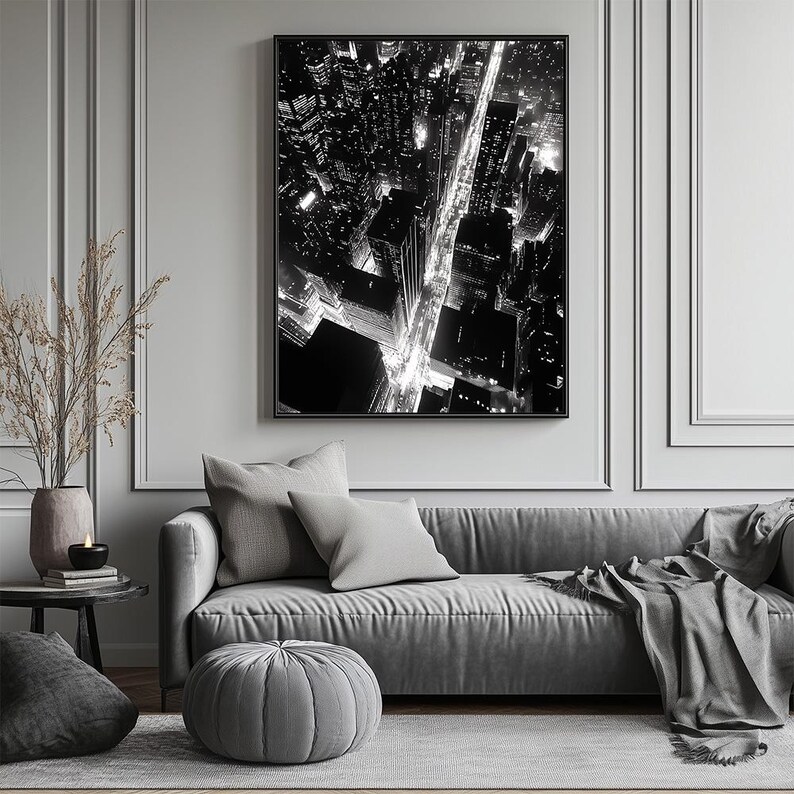 May include: Black and white photograph of a city skyline at night. The image shows a street lined with buildings and streetlights. The photo is framed and hanging on a wall above a gray couch.