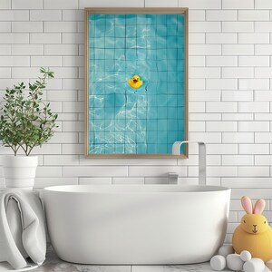 May include: A framed print featuring a yellow rubber duck floating in a blue pool. The print is in a light brown frame and hangs above a white bathtub. A small plant in a white pot and a yellow stuffed bunny are also in the scene.