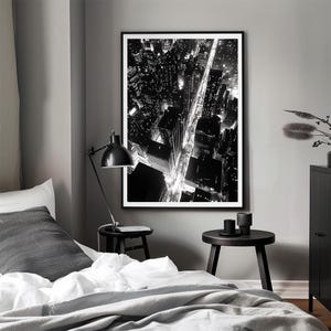 May include: Black and white photograph of a city skyline at night. The photo shows a street lined with buildings and streetlights. The photo is framed and hanging on a wall.