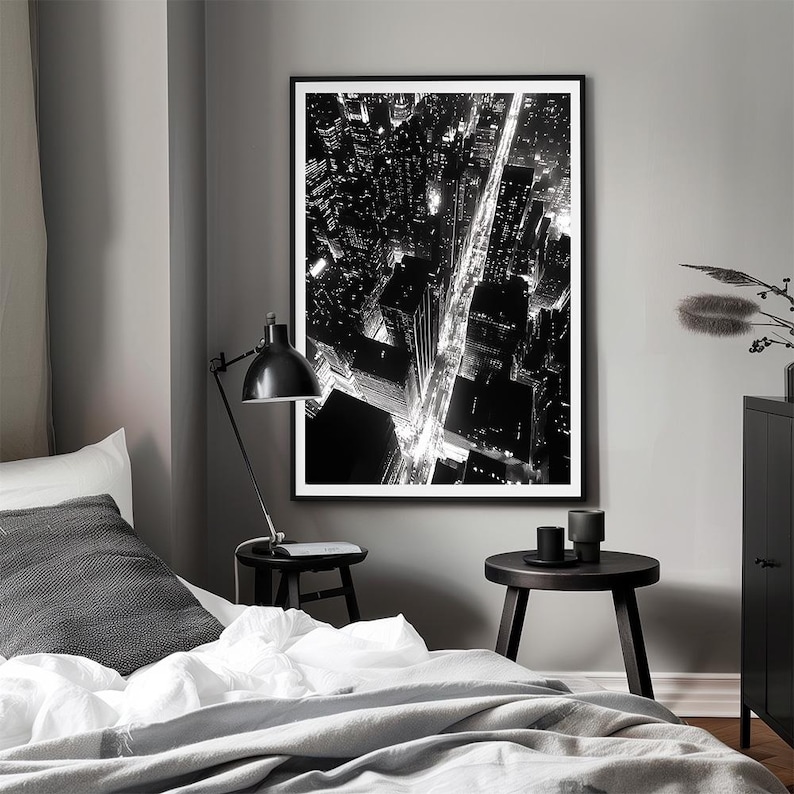 May include: Black and white photograph of a city skyline at night. The photo shows a street lined with buildings and streetlights. The photo is framed and hanging on a wall.