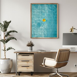 May include: A framed print featuring a top-down view of a swimming pool with blue tiles and a yellow rubber duck floating in the water. The print is in a wooden frame and hangs on a white wall above a wooden desk with a computer and chair.