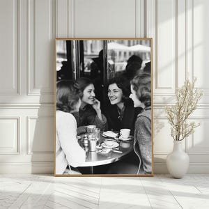 Op de afbeelding: Drie vrouwen zitten aan een tafel in een café, lachen en praten. Ze genieten van koffie en gebak. Het café is ingericht in een vintage stijl.