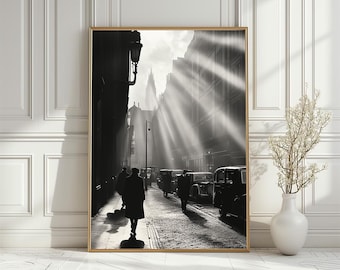 May include: Black and white photograph of a city street with people walking and cars driving. The sun is shining brightly and the light is streaming through the buildings. The photo is taken from a low angle, looking up at the buildings.