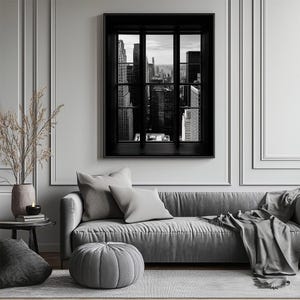 K&ouml;nnte beinhalten: Schwarz-Wei&szlig;-Fotografie einer Stadtlandschaft, die durch ein Fenster mit schwarzem Rahmen betrachtet wird. Das Fenster befindet sich an einer wei&szlig;en Wand &uuml;ber einem grauen Sofa mit Kissen und einem grauen Ottoman. Eine Vase mit getrockneten Pflanzen steht auf einem Beistelltisch.
