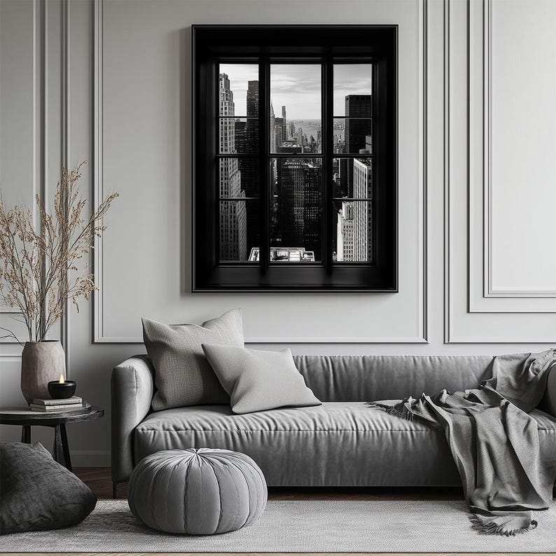 K&ouml;nnte beinhalten: Schwarz-Wei&szlig;-Fotografie einer Stadtlandschaft, die durch ein Fenster mit schwarzem Rahmen betrachtet wird. Das Fenster befindet sich an einer wei&szlig;en Wand &uuml;ber einem grauen Sofa mit Kissen und einem grauen Ottoman. Eine Vase mit getrockneten Pflanzen steht auf einem Beistelltisch.