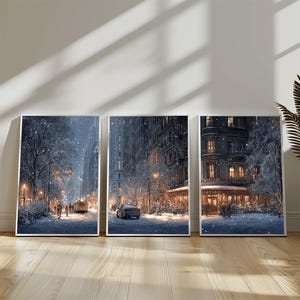 May include: Three framed art prints showing a snowy city scene. The artwork depicts buildings, streetlights, and vehicles on a snow-covered street. The prints are displayed on a wooden floor.
