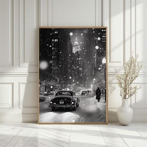 以下が含まれることがあります： 雪が降る街の道を走るビンテージカーの白黒写真。車は、大きな装飾的なグリルと左右に1つずつヘッドライトが付いたクラシックなセダン。通りは背の高い建物が立ち並び、雪が激しく降っている。写真はビンテージ感があり、雪降る街の美しい風景を捉えている。