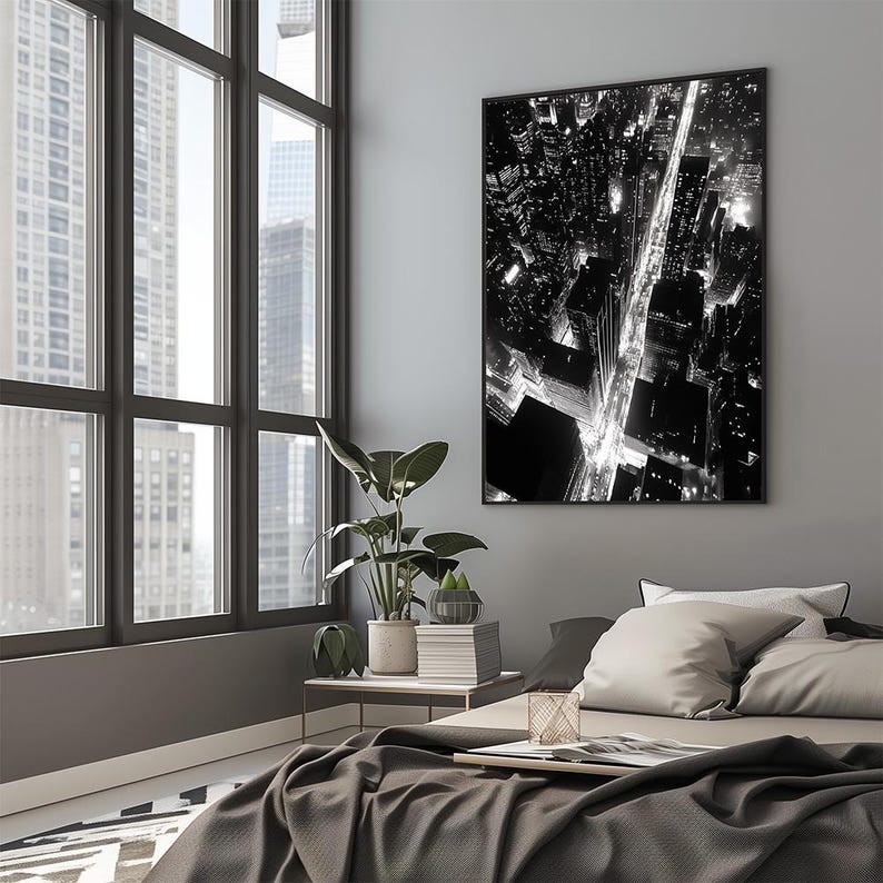 May include: A black and white photograph of a city skyline at night. The photo shows a street with buildings and streetlights on either side. The photo is framed in a black frame and hanging on a gray wall.