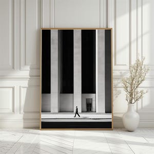 Puede incluir: Impresión artística enmarcada con una fotografía arquitectónica en blanco y negro. La imagen muestra a una persona caminando frente a un edificio con rayas verticales en blanco y negro. La impresión está en un marco dorado y se exhibe contra una pared blanca.