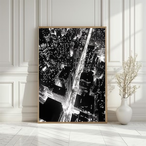 May include: Black and white aerial photograph of a city at night. The photo shows a grid of streets with buildings and lights.