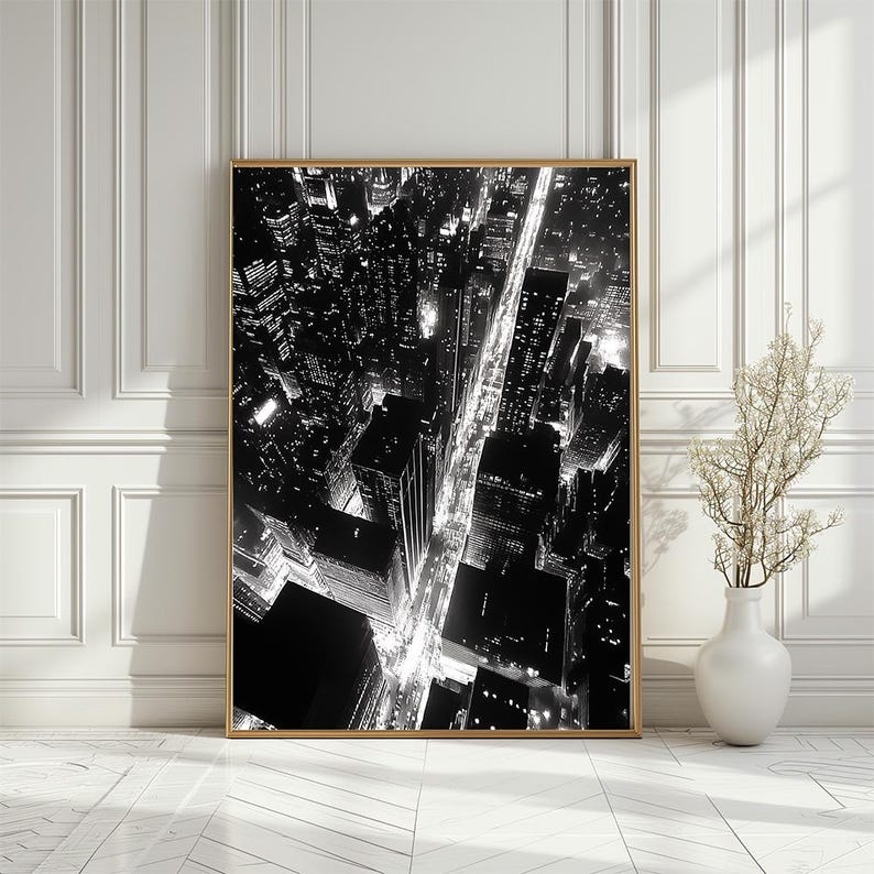 May include: Black and white aerial photograph of a city at night. The photo shows a grid of streets with buildings and lights.